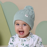 Baby wearing a light green knit hat with a pom-pom against a beige and green heart-shaped background